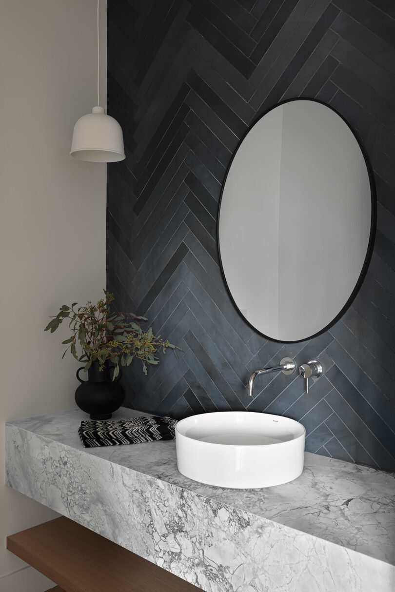 Bathroom with a round mirror, herringbone tile wall, white hanging light, marble countertop, round white sink, and black vase with greenery.