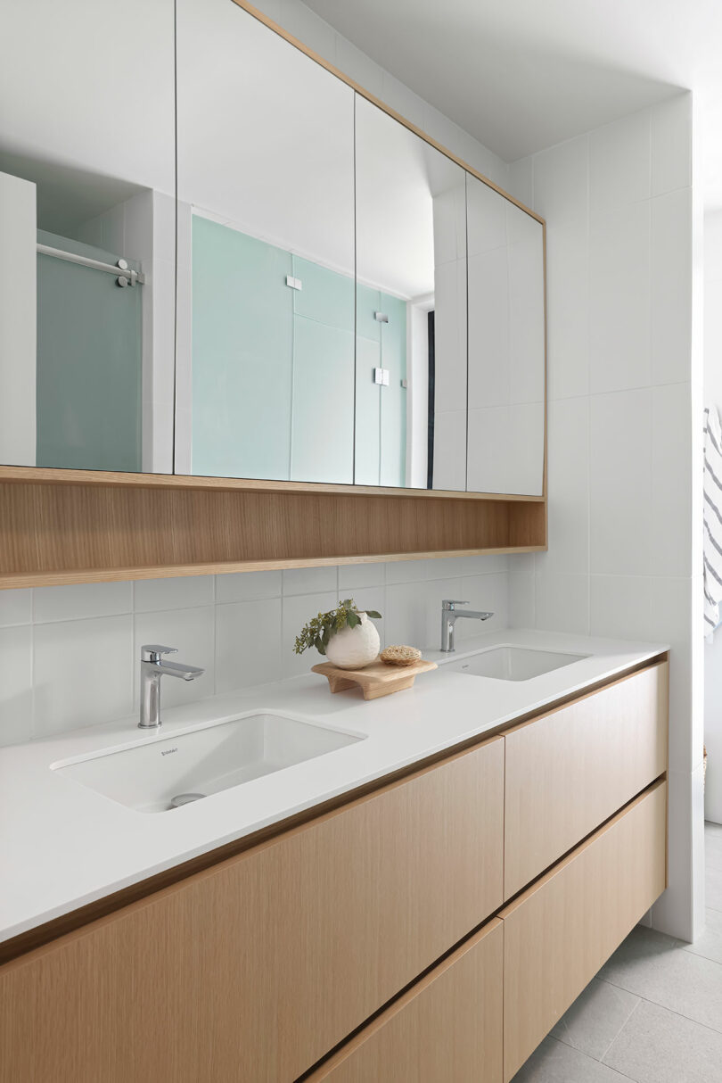 Modern bathroom with a double sink vanity, wooden cabinets, large mirrors, and a decorative plant. The flooring is light gray, and the walls are tiled in white.