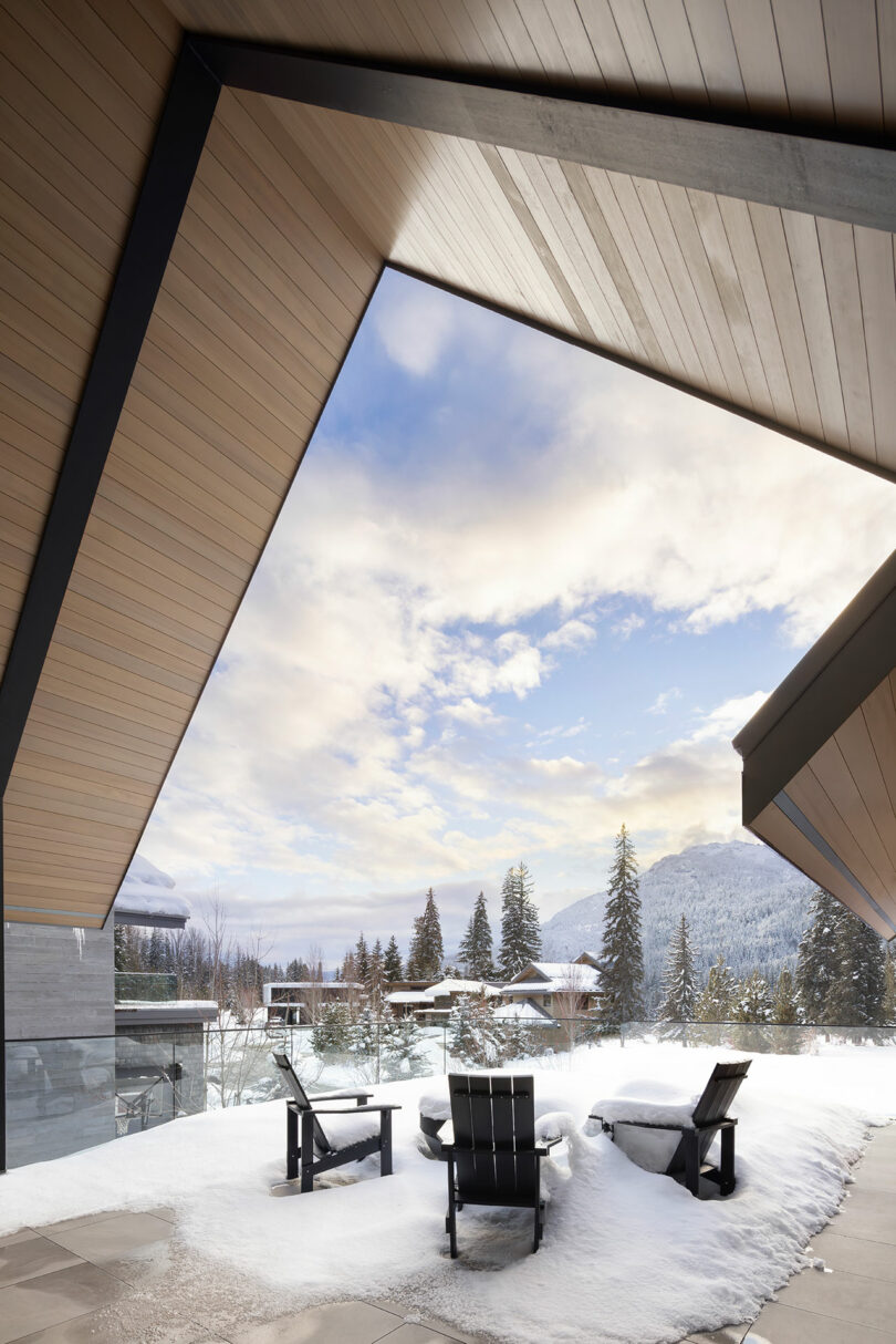 Snowy patio with three empty chairs under a modern wooden roof. Snow-covered trees and mountains are visible in the background under a partly cloudy sky.