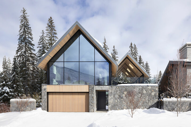 Modern house with large glass windows, stone walls, and wood accents, surrounded by snow and evergreen trees.