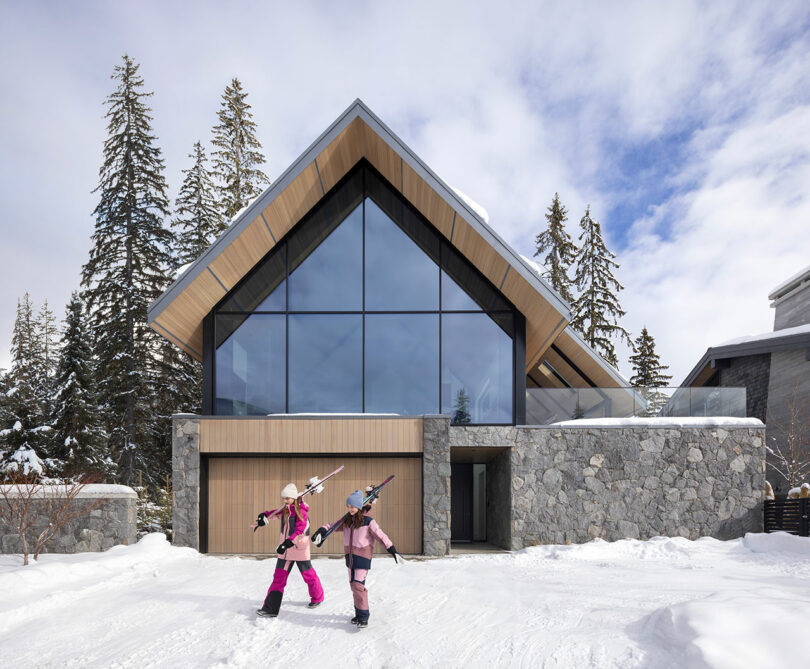 Two people in winter clothing carry skis in front of a modern A-frame house with large glass windows, surrounded by snow and tall trees.