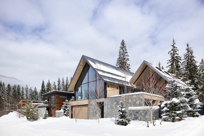 Modern house with large windows and stone facade surrounded by snow and pine trees under a cloudy sky.