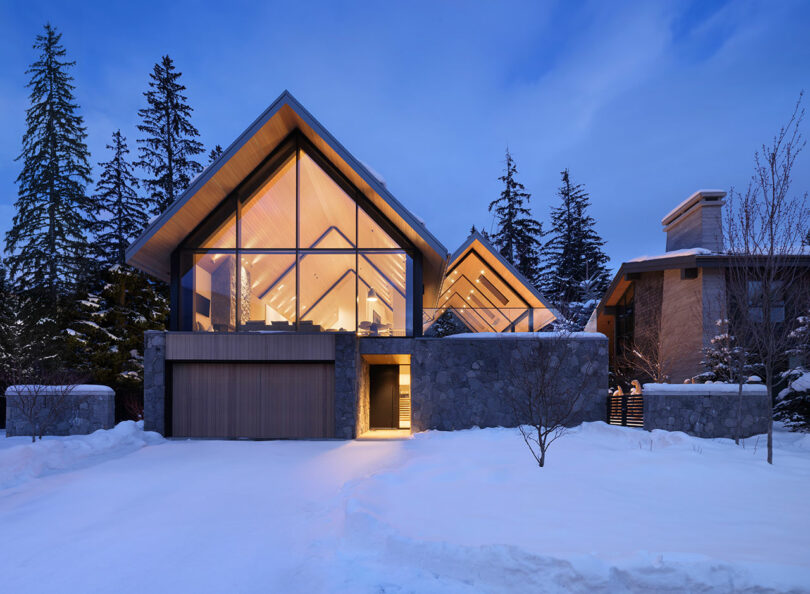 A modern house with large glass windows and a sloped roof is surrounded by snow and tall trees under a twilight sky. The interior lighting gives the house a warm glow.