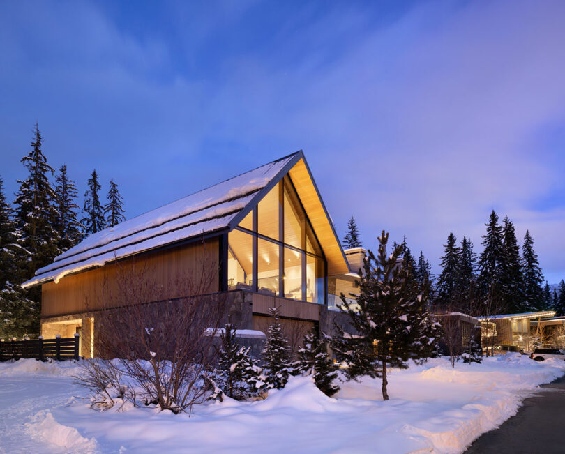 A modern, snow-covered house with large glass windows and a steep roof, surrounded by pine trees during twilight.