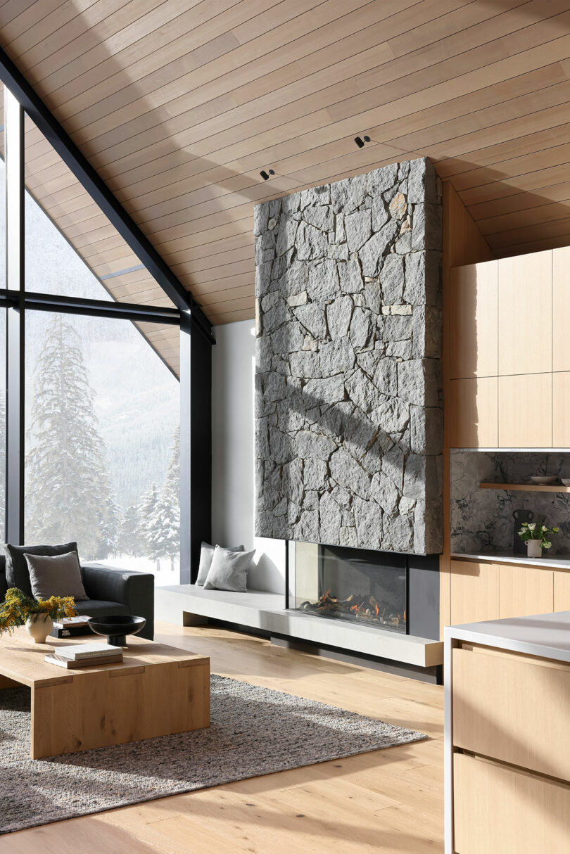 Modern living room with a large stone fireplace, wooden ceiling, and large windows showcasing a snowy landscape. Neutral-toned furniture and decor complete the space.