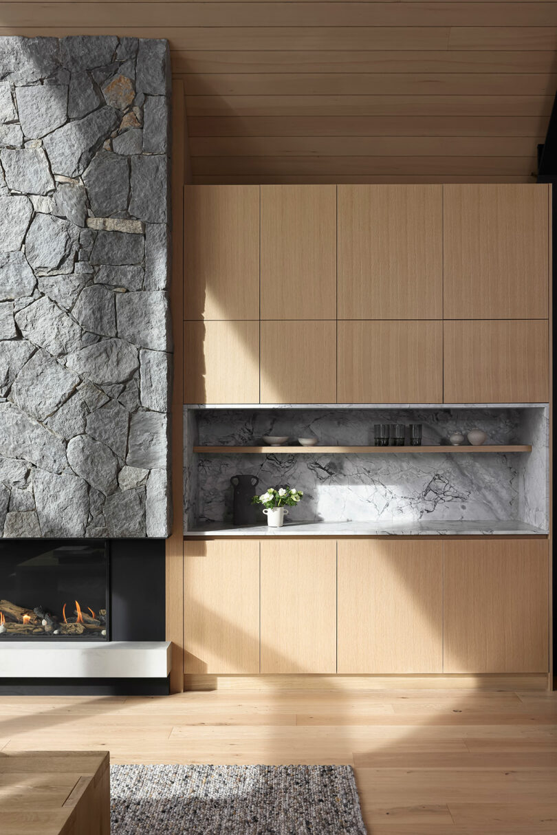 Modern living room with a stone fireplace and light wood cabinetry. A recessed shelf with a small plant and decor items is in the center. Light wood flooring and a woven rug complete the space.