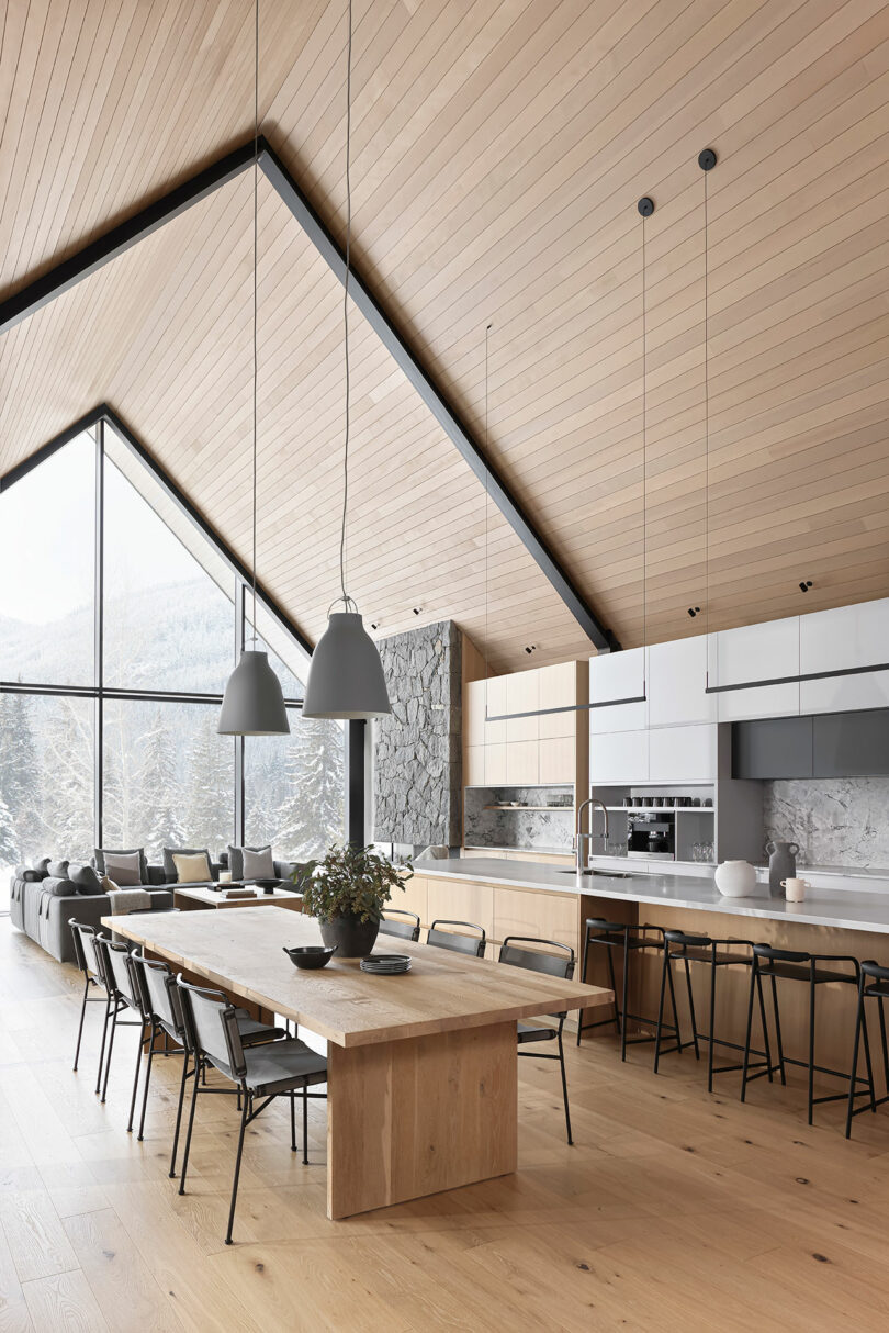 Spacious modern kitchen and dining area with wooden ceiling and floor, large windows, stone wall, long dining table, and hanging pendant lights.