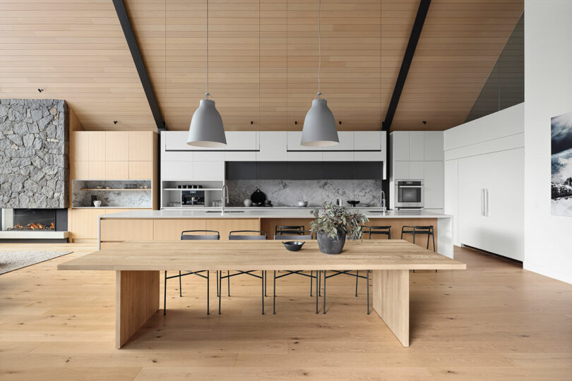 Modern kitchen with a large wooden dining table, black chairs, pendant lights, and a stone fireplace. White cabinets and marble backsplash enhance the sleek design. .