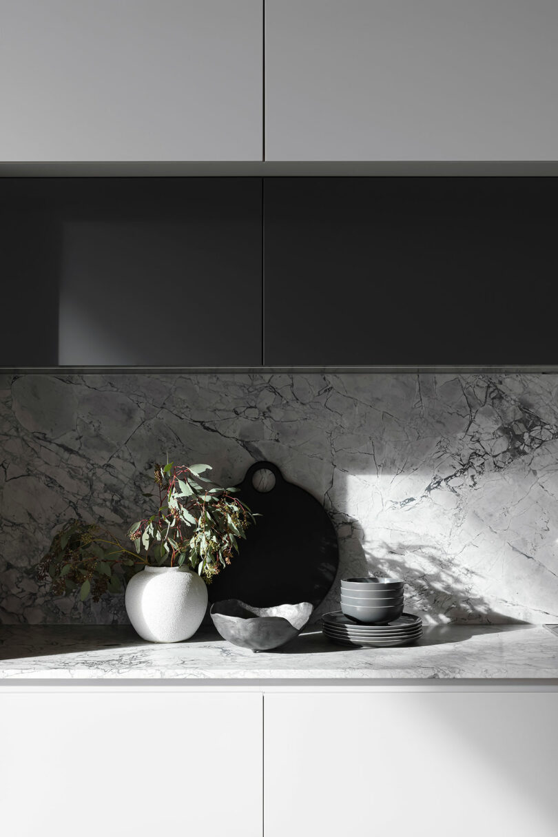 Minimalist kitchen countertop with a white vase holding branches, a dark cutting board, stacked bowls, and a dark bowl, set against a marble backsplash and under dark cabinets.