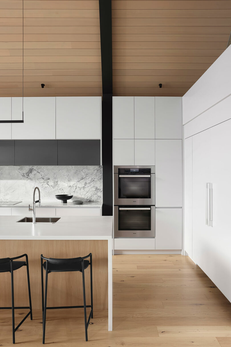 Modern kitchen with white cabinetry, a marble backsplash, and wood paneling. Features a kitchen island with sink and black stools, and built-in ovens.