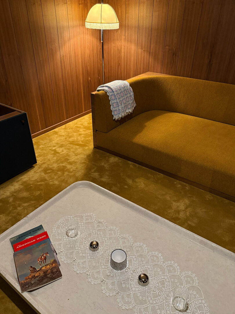 A retro living room with a mustard sofa, wooden walls, and a lamp. A coffee table with a lace runner, book, and candles sits on a mustard carpet.