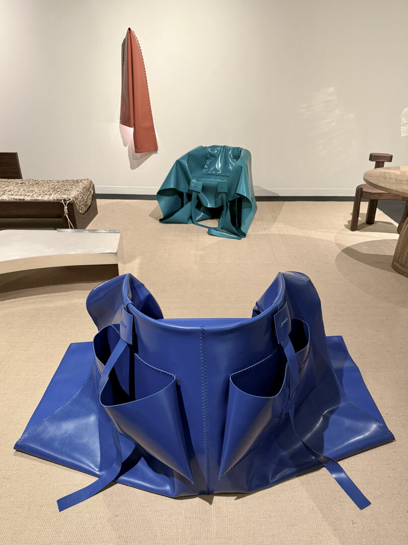 Installation view with blue, teal, and brown abstract sculptural objects arranged on a neutral floor in a minimalist gallery space.