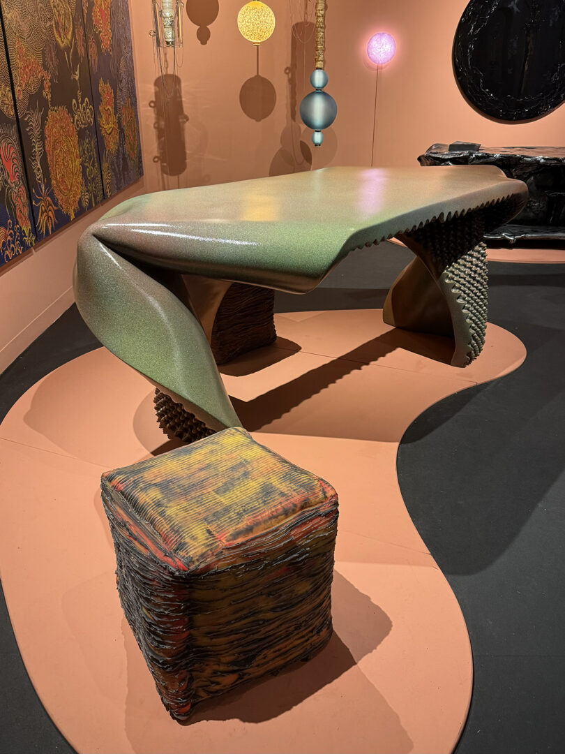 A modern, sculptural table with a twisted design and a textured surface, paired with a cube-shaped stool on a decorative floor. Colorful abstract art hangs on the wall behind.