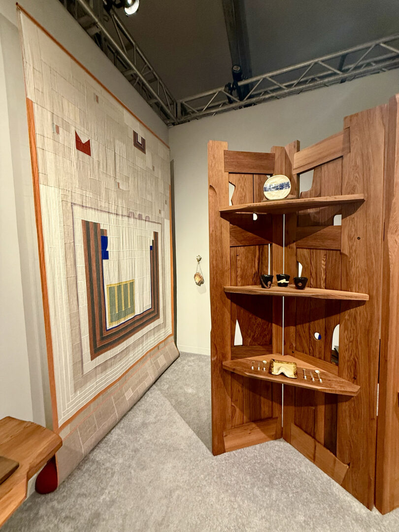 A room with a geometric tapestry on the wall and a wooden shelf displaying pottery and small sculptures.