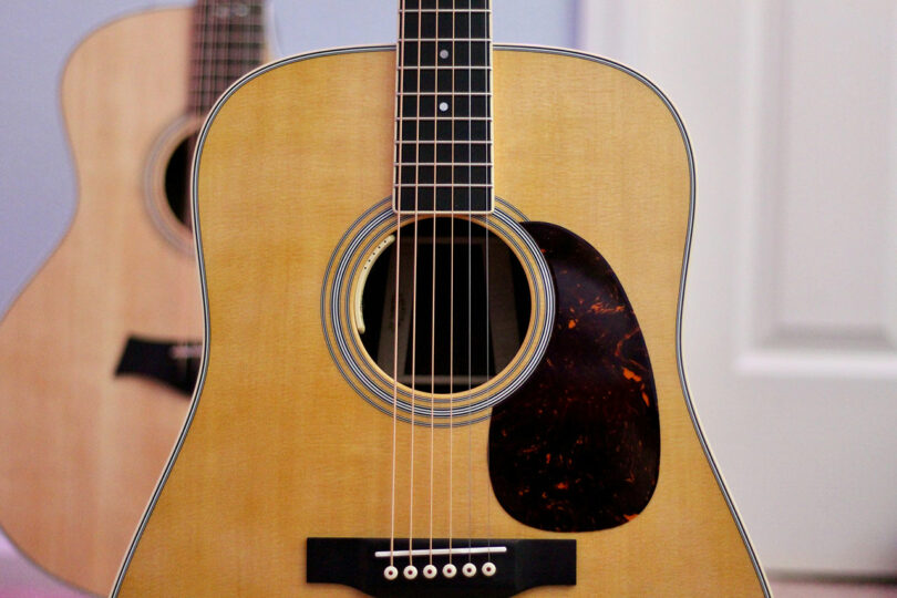 Close-up of a wooden acoustic guitar with a tortoiseshell pickguard, reminiscent of the craftsmanship associated with Dan Harden. Another guitar is partially visible in the background.