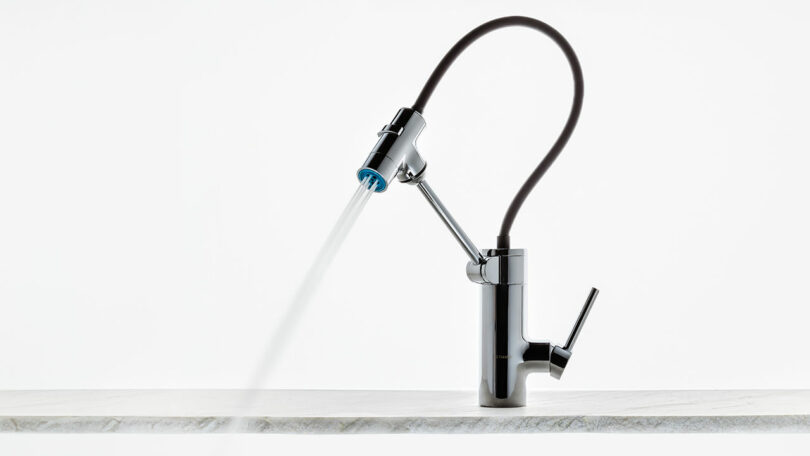 Designed by Dan Harden, a modern chrome kitchen faucet with a flexible black hose is mounted on the countertop. It elegantly pours a stream of water.