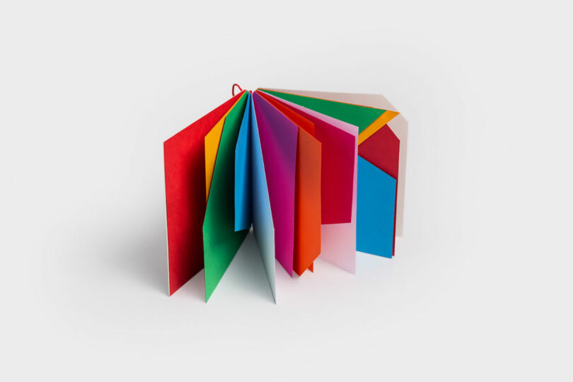 A fan of colorful, overlapping paper sheets designed with Theodora Alfredsdottir's unique touch, displayed in an accordion fold against a plain background.