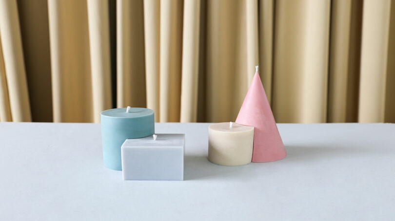 Four candles in geometric shapes and pastel colors, including blue, gray, cream, and pink, designed by Theodora Alfredsdottir, are elegantly placed on a light surface with beige curtains gently draping in the background.