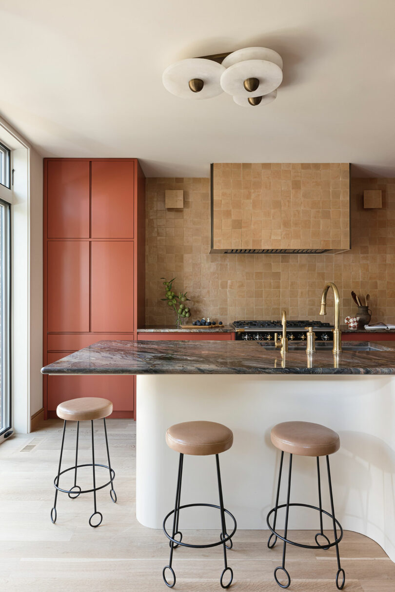 Modern kitchen with a large marble island, three stools, a brass faucet, and red cabinetry. Beige tiles cover the backsplash, and a contemporary ceiling light fixture is visible.