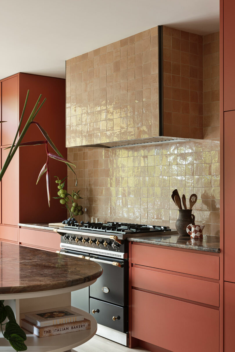 A kitchen with terra-cotta cabinets, a black oven with multiple knobs, a tiled backsplash, and a countertop with a vase holding a large flower. Cooking utensils are placed in a cup next to the oven.