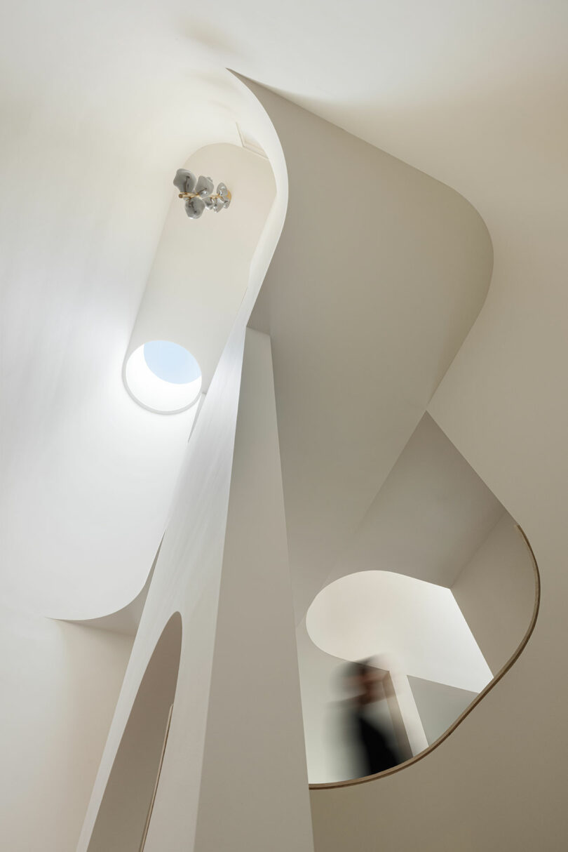 A person walks up a staircase in a modern white-walled building with curved architectural elements and a skylight.