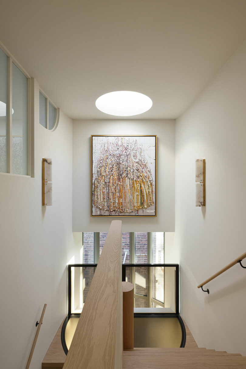 A modern white staircase with wooden handrails, featuring a large abstract painting and a round ceiling light overhead.