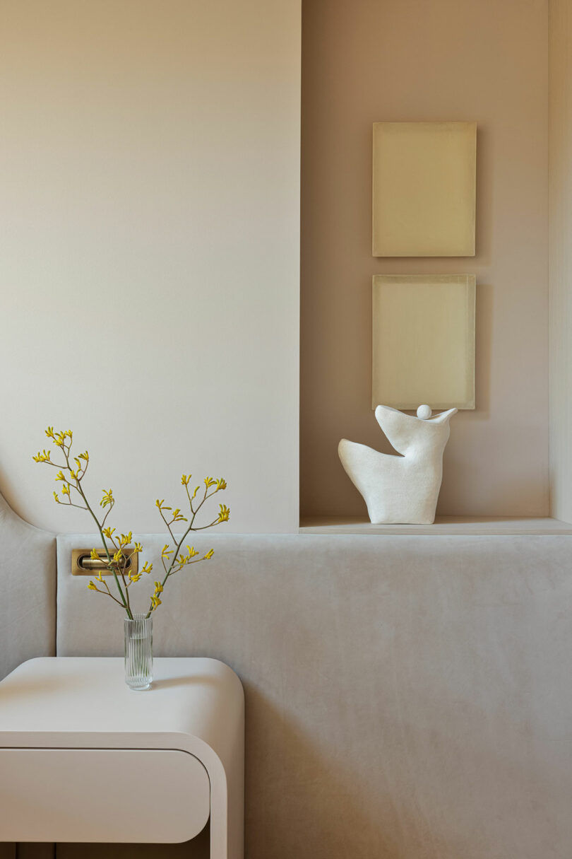 A minimalist interior with a white sculptural piece, two beige rectangular wall art pieces, and a small vase with yellow flowers on a white table.