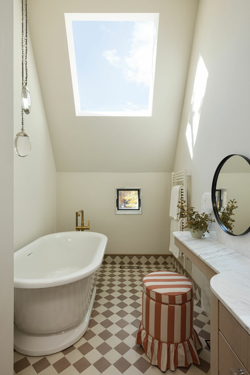 A bathroom with a white freestanding bathtub, a small window, a round mirror, and a patterned beige floor. A striped stool is in front of a vanity, and a large skylight brightens the room.