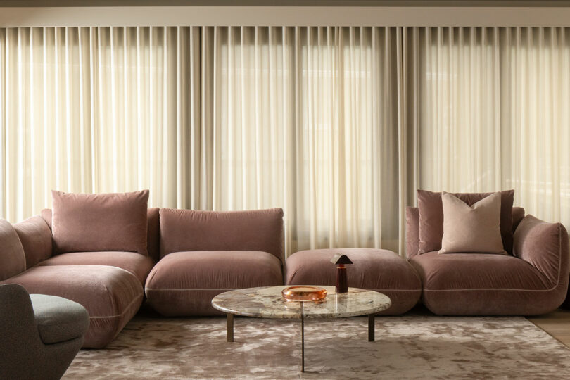 A modern living room with a pink sectional sofa, a round marble coffee table, and light-colored curtains.