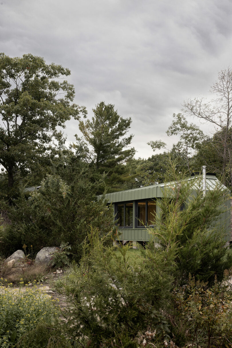 A small green house surrounded by dense trees and bushes under a cloudy sky.