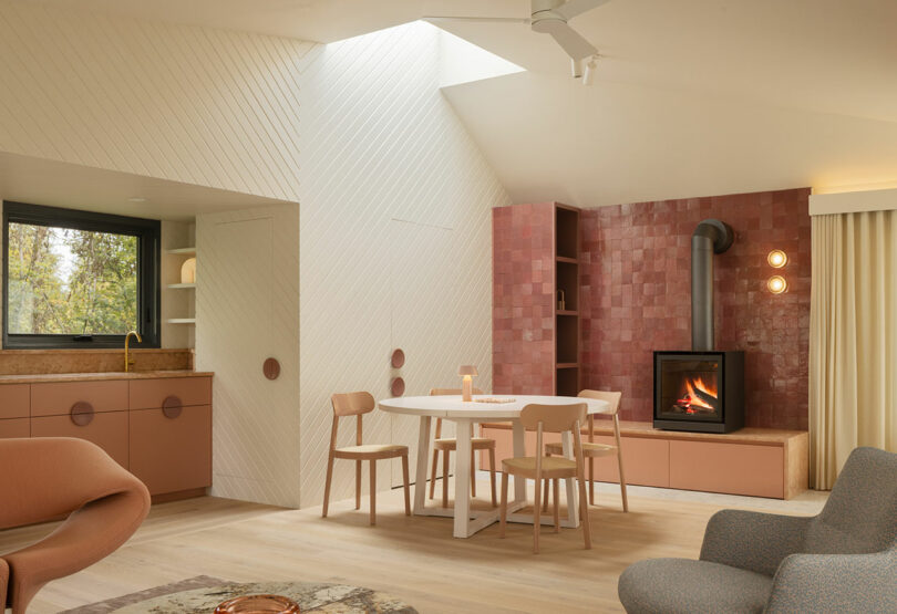 Modern living room with a cozy fireplace, light wood floors, and earth-toned furniture. A dining table with chairs is near a skylight, and the kitchen has pink cabinets.