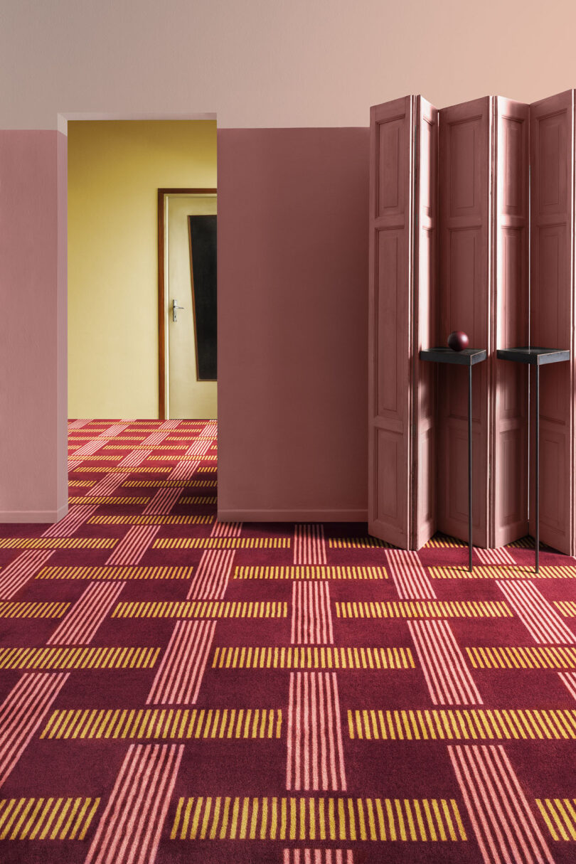 Room with patterned red and yellow carpet, pink walls, a folding screen, and visible doorway leading to another room with yellow walls and a door.