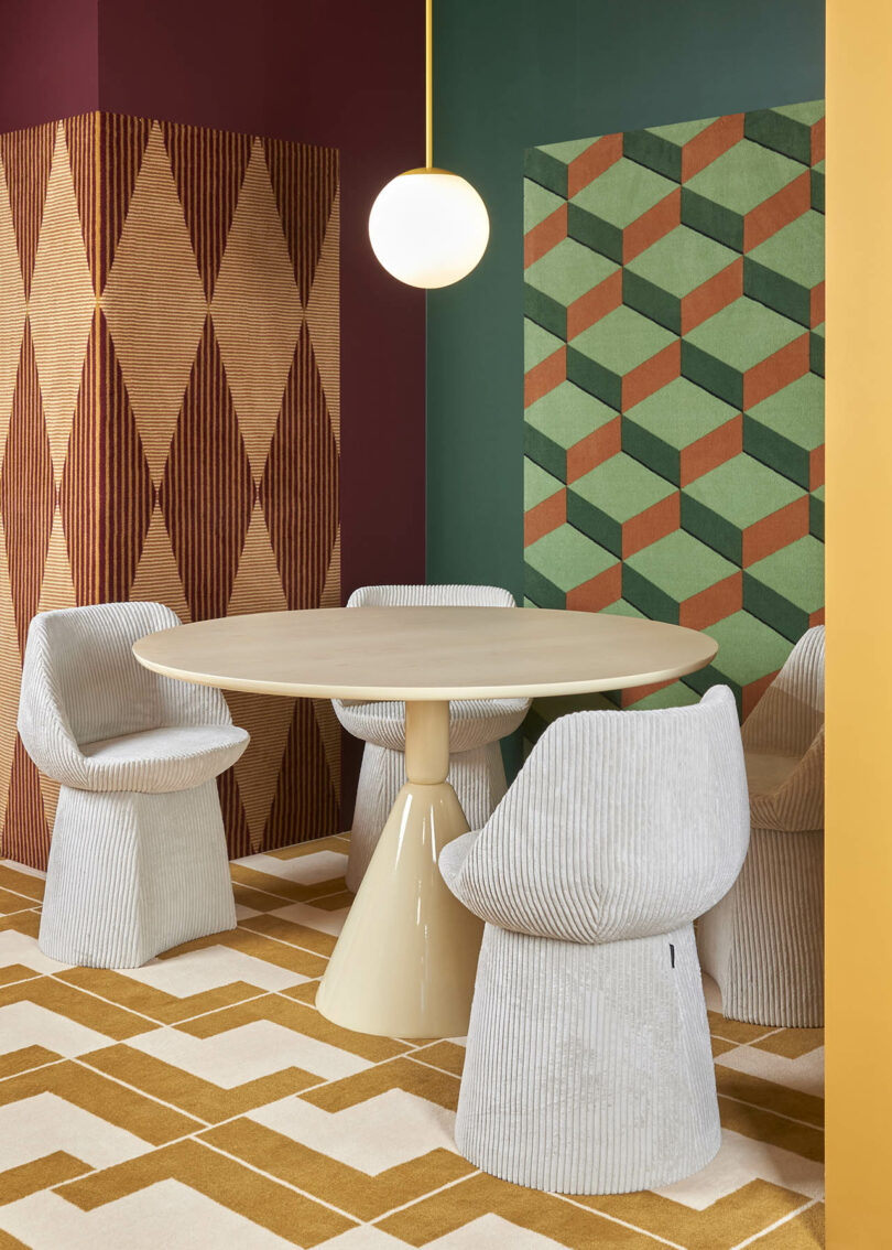 Modern dining area with a round beige table, four textured white chairs, and geometric patterned walls and floor in green, yellow, and red tones. A spherical light fixture hangs overhead.