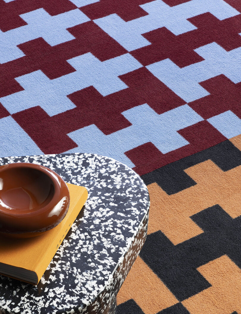 Geometric pattern rug with red, blue, and tan shapes. A speckled black and white table holds a brown bowl and yellow book.