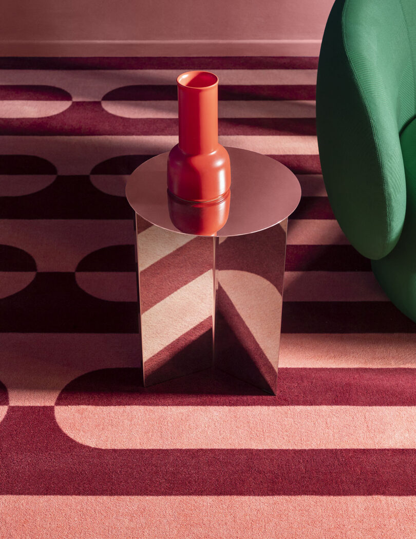 A red vase on a mirrored side table sits atop a patterned carpet with geometric designs in shades of red and pink. A green chair is partially visible on the right.