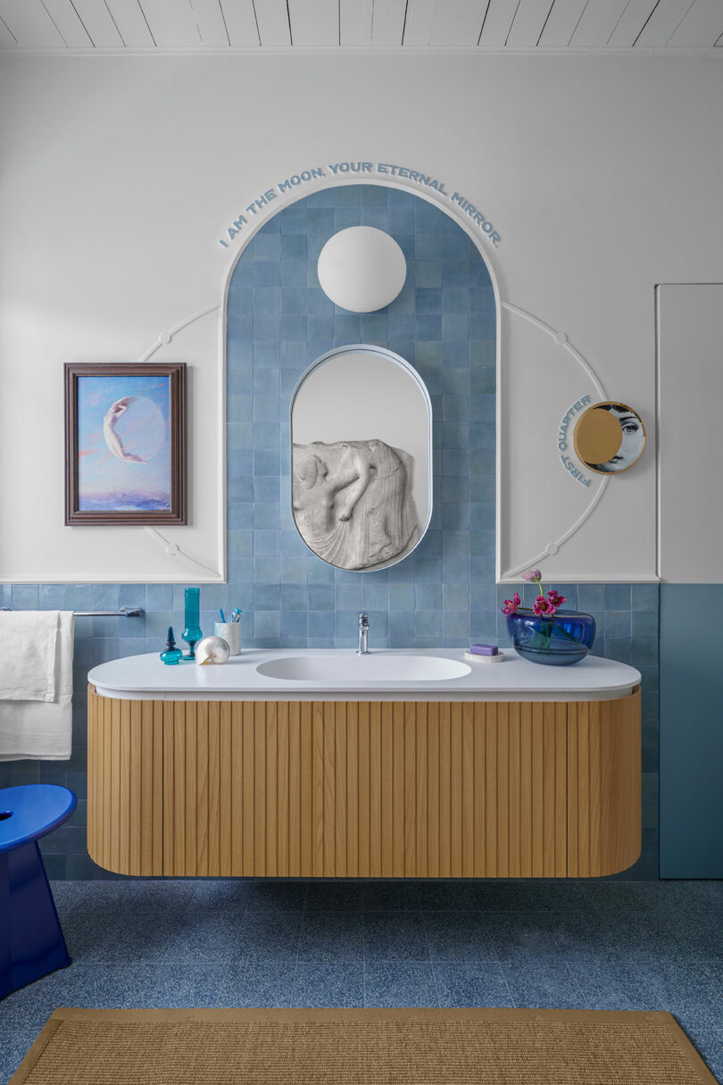 Modern bathroom with a curved wooden vanity, blue tiles, and a round mirror. Wall inscription reads, "I am the moon, your eternal mirror." Decor includes artwork and a small mirror.