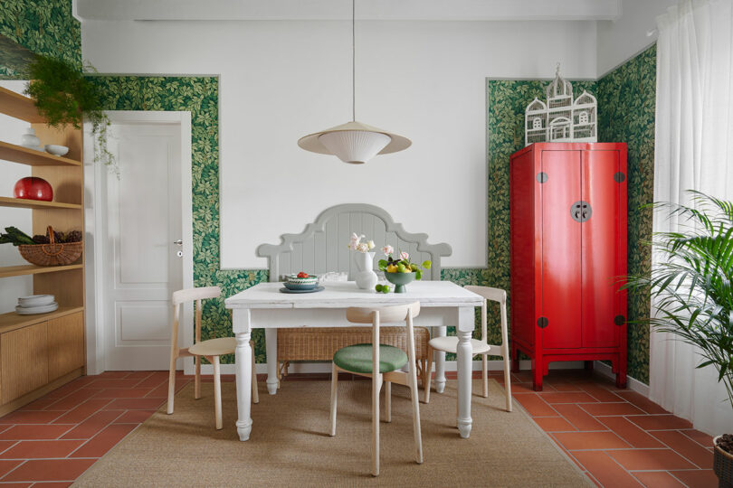 A dining room with a white table, green chairs, red cabinet, and green patterned wallpaper. A pendant light hangs above the table, and a woven rug is on the brick-tile floor.