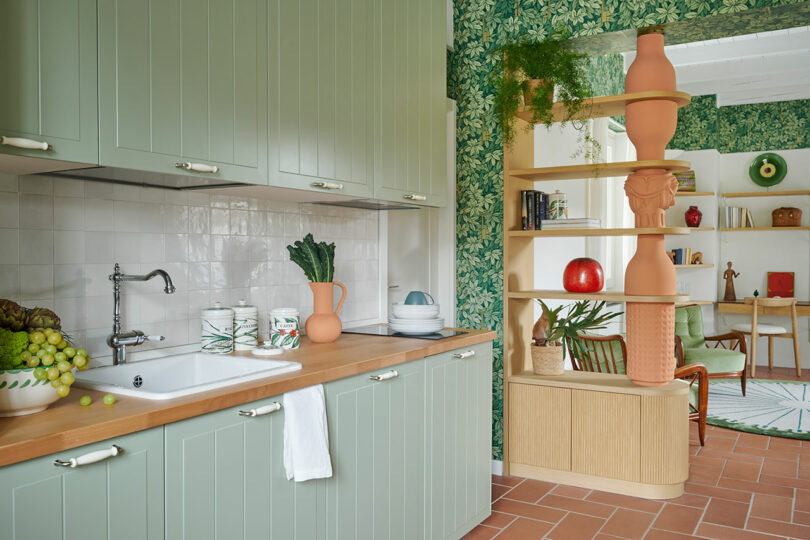 A kitchen with green cabinets, a wooden countertop, a white sink, and a tiled backsplash. A shelf with decorative items is next to a wallpapered wall with a leafy pattern, leading to a cozy room.