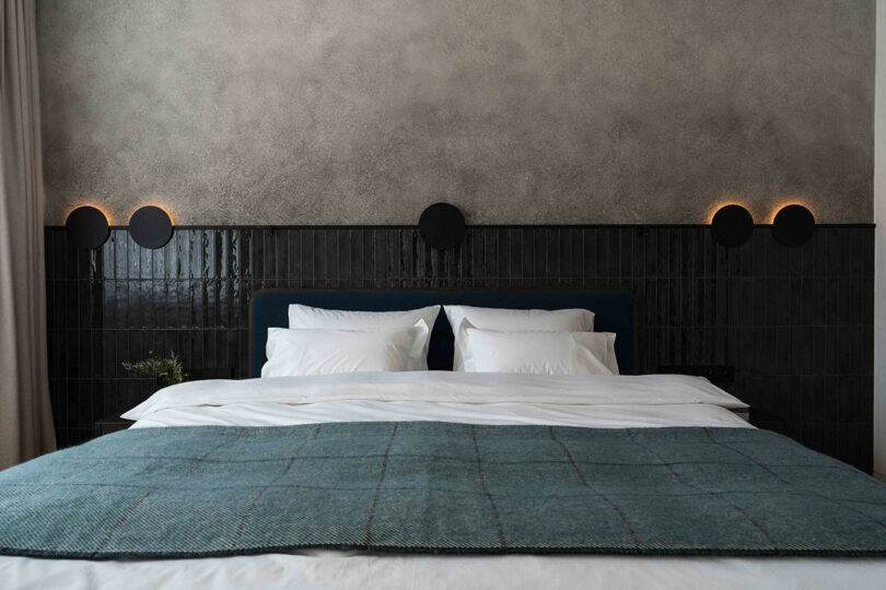 A neatly made bed with a dark headboard against a textured wall. Circular lights are mounted above the headboard, and a blue blanket covers the bed.