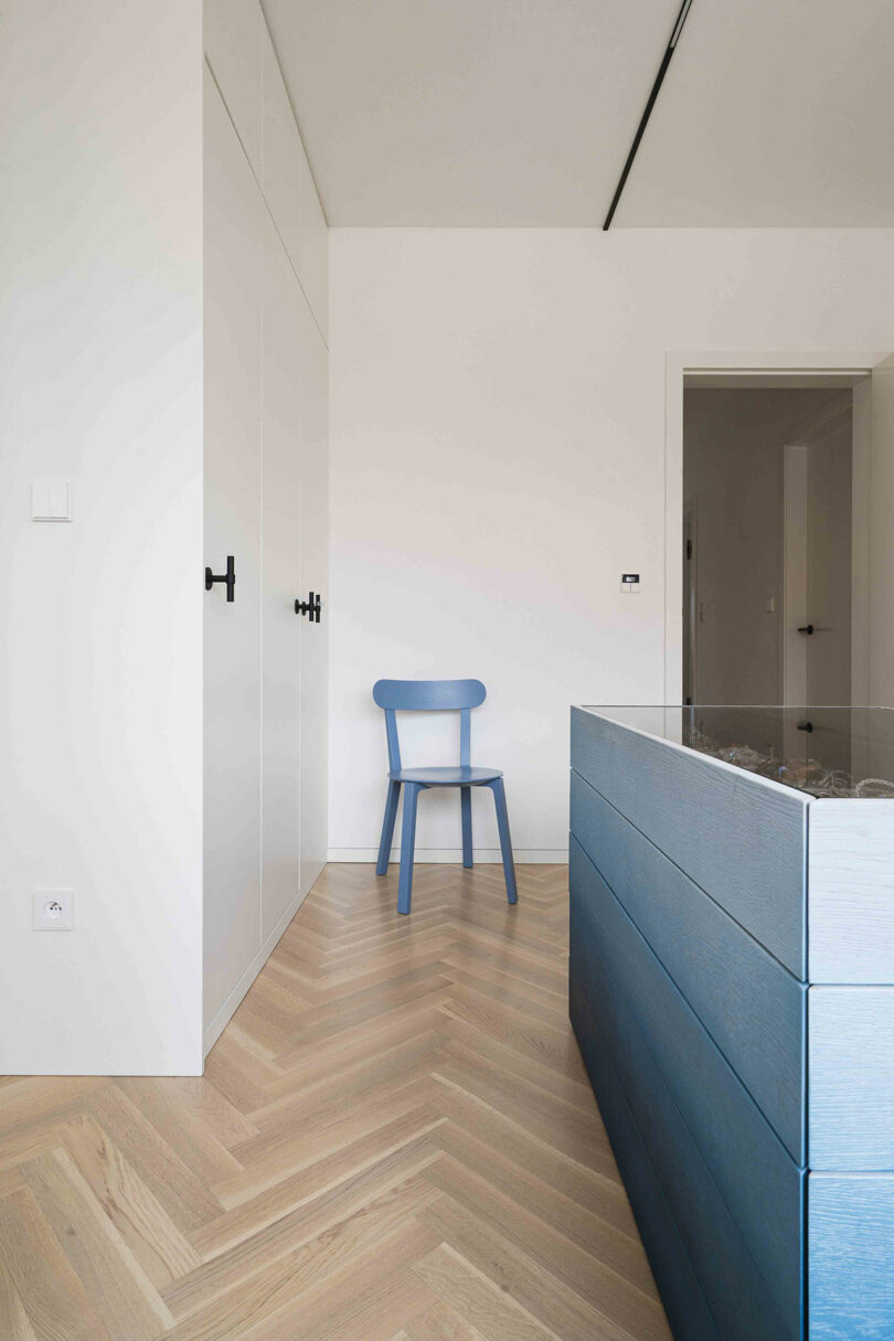 Minimalist room with light wood herringbone floor, a blue chair near a white wall with black handles, and a blue side cabinet.