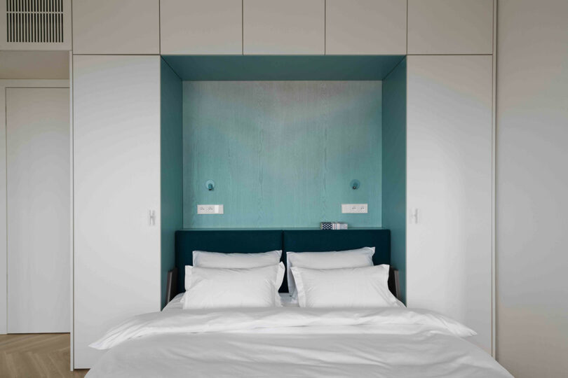 Minimalist bedroom with a double bed, white bedding, and teal accent wall. Flanked by white built-in cupboards, the headboard is dark green, and two wall lights are above it.