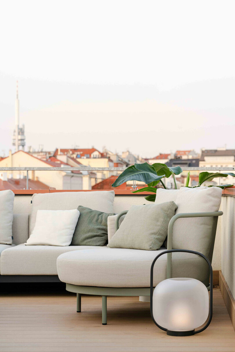 Rooftop terrace with beige and green cushioned seating, a modern lantern, and a view of city rooftops and a tower in the distance.