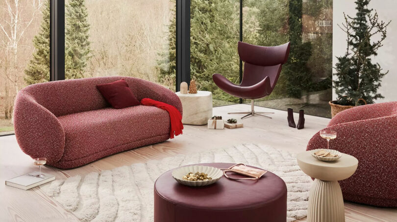 A modern living room with red seating, including a sofa and chair. The room features large windows, a round ottoman, and a small table with snacks. A forest view is visible outside