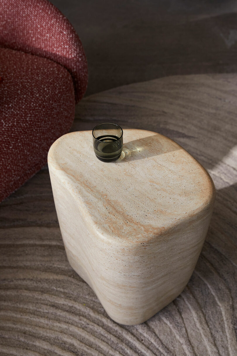 A small dark glass sits on an irregularly shaped beige stone side table. A textured red cushion is partly visible in the background