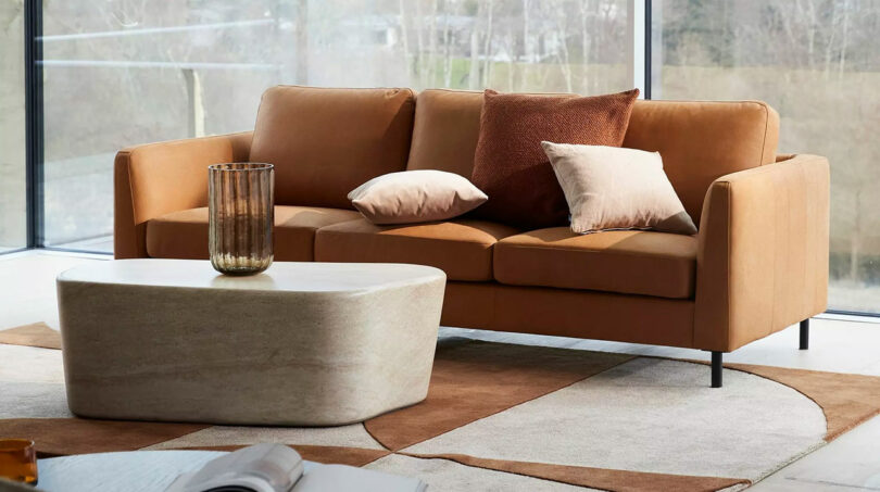 A modern living room with a brown sofa adorned with pillows, a rectangular stone coffee table, and large windows showing a blurred outdoor view