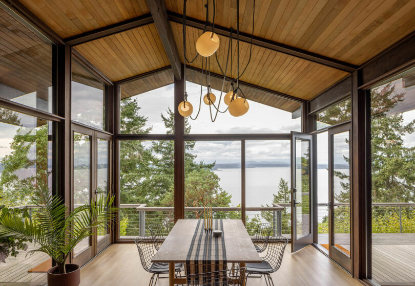 A modern dining area with large windows overlooks a serene lake. A wooden table and wire chairs are centered under unique hanging lights. Lush greenery is visible outside.