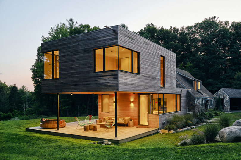 Modern two-story house with large windows, wooden exterior, and an outdoor deck with seating, set in a grassy area with trees in the background.