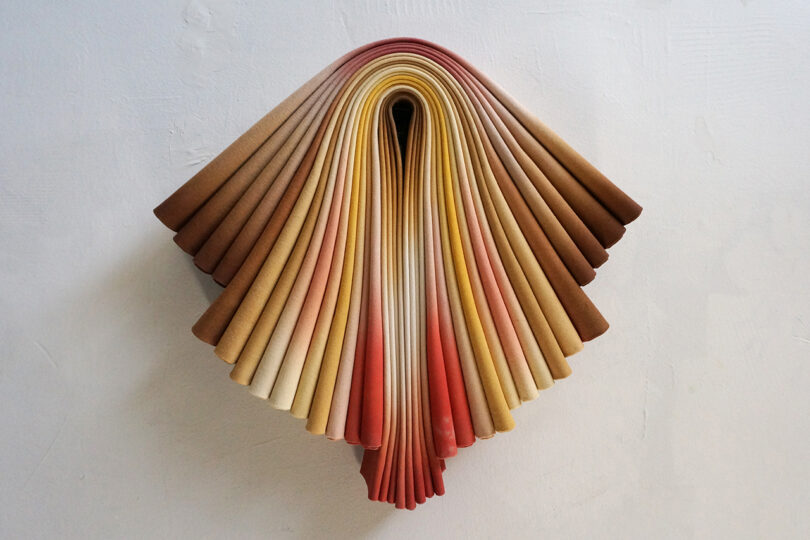 Folded pages resembling a fan with a gradient of earth tones, mounted against a white wall.