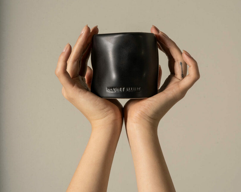 Two hands gently hold a black, square-shaped cup with embossed text, blending seamlessly into the surrounding home furnishings set against a neutral background.