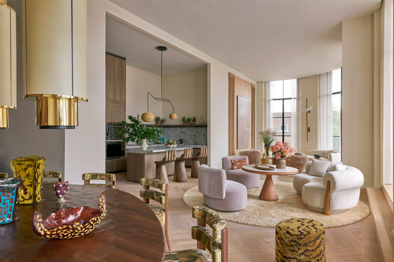 A modern open-plan living and dining area with eclectic decor, soft neutral tones, patterned chairs, round wooden tables, and large windows with sheer curtains.